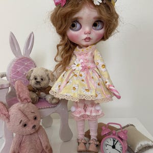 May include: A doll with blonde hair wearing a yellow and white daisy print dress with pink accents. The doll is holding a candy cane and is sitting on a white chair with a pink cushion. There are two teddy bears in the image, one sitting on the chair and one sitting on the floor.