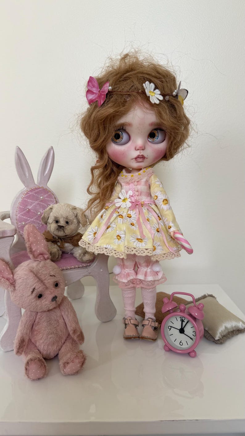 May include: A doll with blonde hair wearing a yellow and white daisy print dress with pink accents. The doll is holding a candy cane and is sitting on a white chair with a pink cushion. There are two teddy bears in the image, one sitting on the chair and one sitting on the floor.