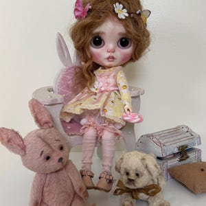 May include: A doll with brown hair wearing a yellow and pink dress sits on a white chair. The doll is holding a pink lollipop. There are two stuffed animals, a pink bunny and a white dog, sitting on the floor in front of the doll.