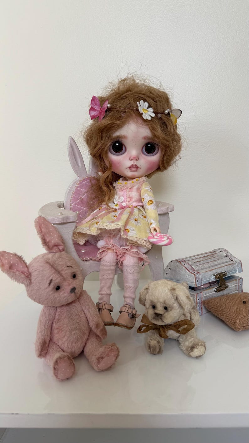 May include: A doll with brown hair wearing a yellow and pink dress sits on a white chair. The doll is holding a pink lollipop. There are two stuffed animals, a pink bunny and a white dog, sitting on the floor in front of the doll.