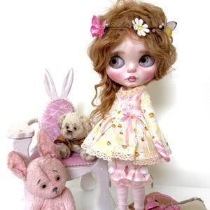May include: A doll with long, curly, light brown hair wearing a yellow and white daisy print dress with pink accents. The doll is holding a pink and white candy cane. The doll is sitting on a white chair with pink upholstery. There are two pink teddy bears in the image, one sitting on the chair and the other sitting on the floor. There is a pink alarm clock on the floor.