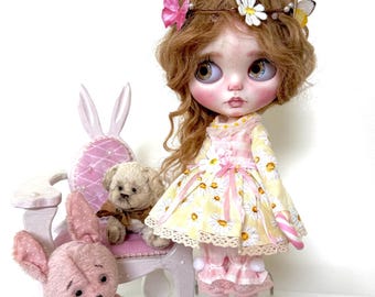 Könnte beinhalten: Eine Puppe mit langen, lockigen, hellbraunen Haaren, die ein gelbes und weißes Kleid mit Gänseblümchenmuster und rosa Akzenten trägt. Die Puppe hält einen rosa und weißen Zuckerstock. Die Puppe sitzt auf einem weißen Stuhl mit rosa Polsterung. Auf dem Bild sind zwei rosa Teddybären zu sehen, einer sitzt auf dem Stuhl und der andere auf dem Boden. Auf dem Boden steht ein rosa Wecker.