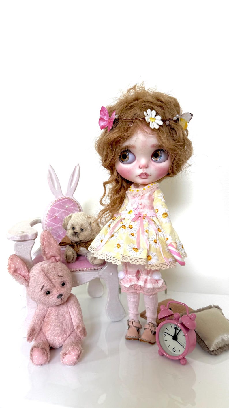May include: A doll with long, curly, light brown hair wearing a yellow and white daisy print dress with pink accents. The doll is holding a pink and white candy cane. The doll is sitting on a white chair with pink upholstery. There are two pink teddy bears in the image, one sitting on the chair and the other sitting on the floor. There is a pink alarm clock on the floor.