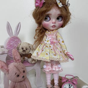 May include: A doll with long brown hair wearing a yellow and white daisy print dress with pink accents, holding a candy cane. The doll is sitting on a white chair with a pink cushion. There are two pink teddy bears and a pink alarm clock in the image.