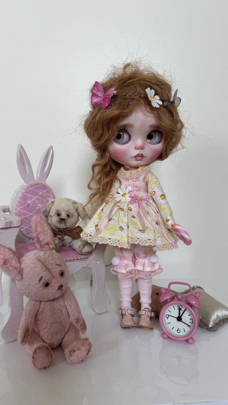 May include: A doll with long brown hair wearing a yellow and white daisy print dress with pink accents, holding a candy cane. The doll is sitting on a white chair with a pink cushion. There are two pink teddy bears and a pink alarm clock in the image.