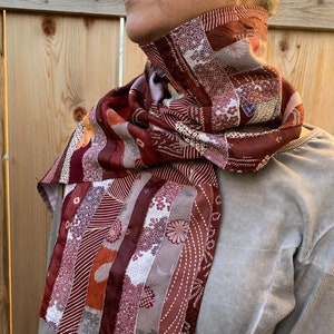 May include: A red, brown, and white patterned scarf made from repurposed fabric. The scarf is tied around the neck of a person wearing a gray sweater.