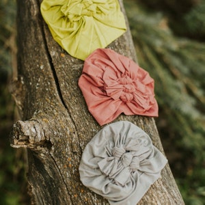May include: Three fabric headbands in different colors: yellow, pink, and gray. Each headband is tied in a knot on top and has a ruffled design.