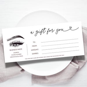 May include: A white gift voucher with black text and a decorative eyelash design. The text reads "a gift for you" and includes fields for recipient, sender, amount, and expiration. The voucher is on a white plate and a light pink cloth.