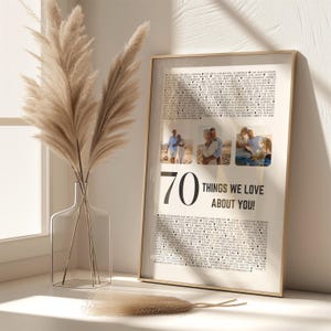 May include: A framed print with the text "70 THINGS WE LOVE ABOUT YOU!" and three photos. The print is in a gold frame and includes a list of text. A clear glass vase with dried pampas grass is in the foreground.