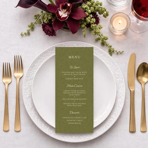May include: A formal table setting featuring a green menu card, white plates, and gold-coloured cutlery. A floral display of dark burgundy calla lilies and orchids is above the plates. A candle, water, and wine glasses complete the scene.
