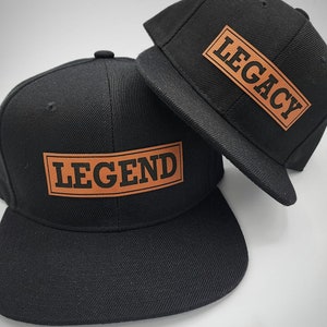 Pode incluir: Dois bonés de beisebol pretos com patches de couro castanho. O boné à esquerda tem a palavra "LEGEND" no patch. O boné à direita tem a palavra "LEGACY" no patch.