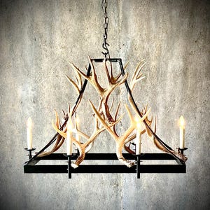 Puede incluir: Una lámpara rústica con astas de ciervo y cuatro luces tipo vela. La lámpara tiene una estructura de metal negro y está suspendida de una cadena. Las astas son de color beige natural, añadiendo un toque de naturaleza al diseño.