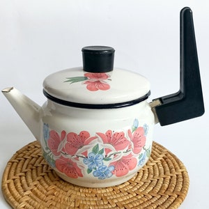May include: White enamel teapot with a black handle and floral design. The teapot is sitting on a woven wicker coaster.
