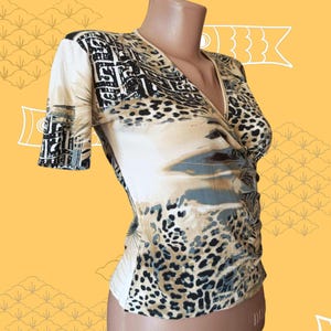 May include: A beige wrap-style top with short sleeves. The top features a mix of patterns, including leopard print, geometric shapes, and abstract designs in black, brown, and grey. The neckline is a V-shape.