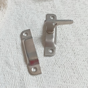 May include: Two silver-colored metal latches. One latch has a lever and is rectangular with rounded edges. The other latch is a rectangular piece with rounded edges. Both latches have holes for screws.