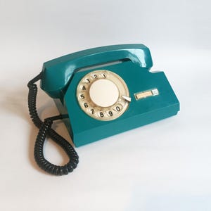 May include: Teal vintage rotary phone with a black coiled cord. The phone features a circular dial with numbers and a cream-colored center. The handset rests on the base, and the design is a classic retro style.