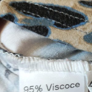 May include: Close-up of patterned fabric in black, beige, and blue. A white label shows "95% Viscose 5% Elastan". The fabric is likely part of a piece of clothing.