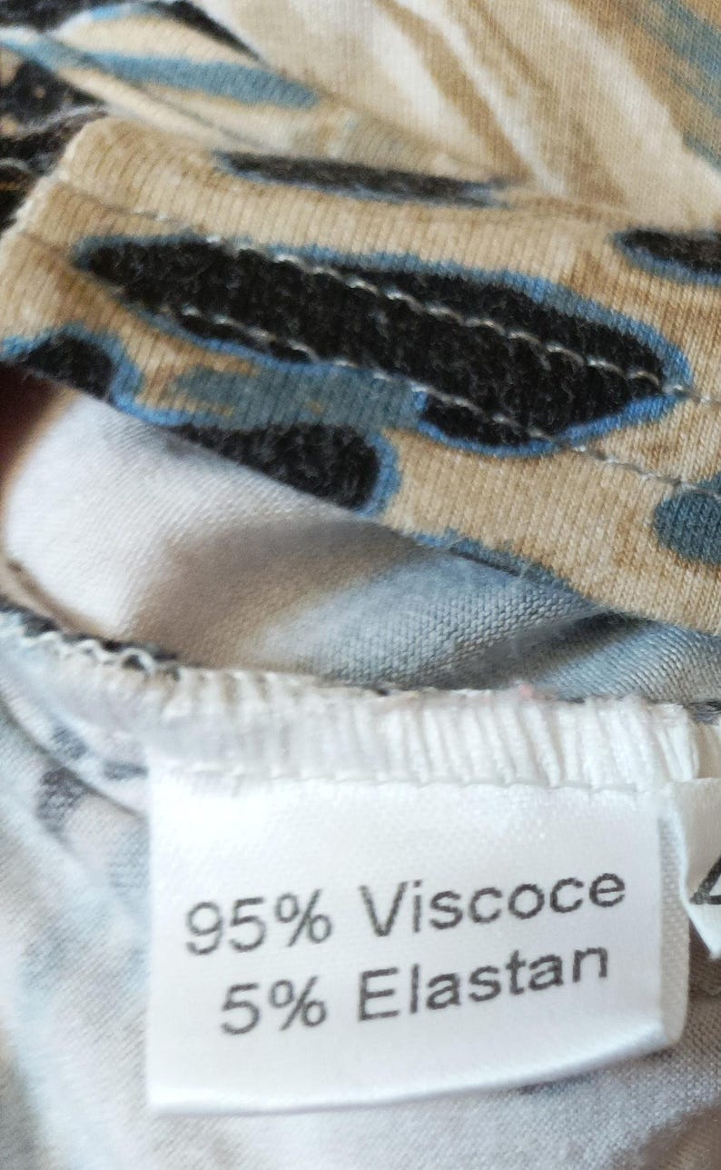 May include: Close-up of patterned fabric in black, beige, and blue. A white label shows "95% Viscose 5% Elastan". The fabric is likely part of a piece of clothing.