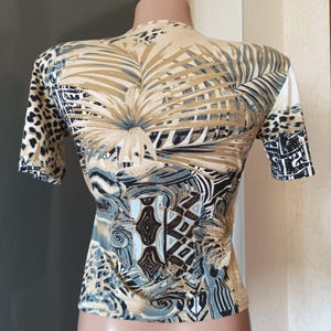 May include: A fitted short-sleeved top with a striking patterned design. The top has a beige base with a large palm leaf design in brown and grey tones. The sleeves feature a leopard print and geometric patterns.