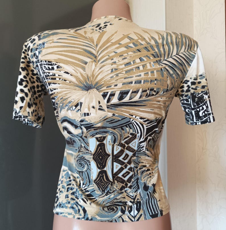 May include: A fitted short-sleeved top with a striking patterned design. The top has a beige base with a large palm leaf design in brown and grey tones. The sleeves feature a leopard print and geometric patterns.