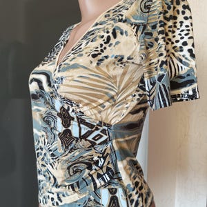 May include: A patterned top with a V-neck and short sleeves. The design features a mix of animal print and abstract shapes in beige, black, and blue. The fabric has a soft appearance. The top has a wrap-style design.