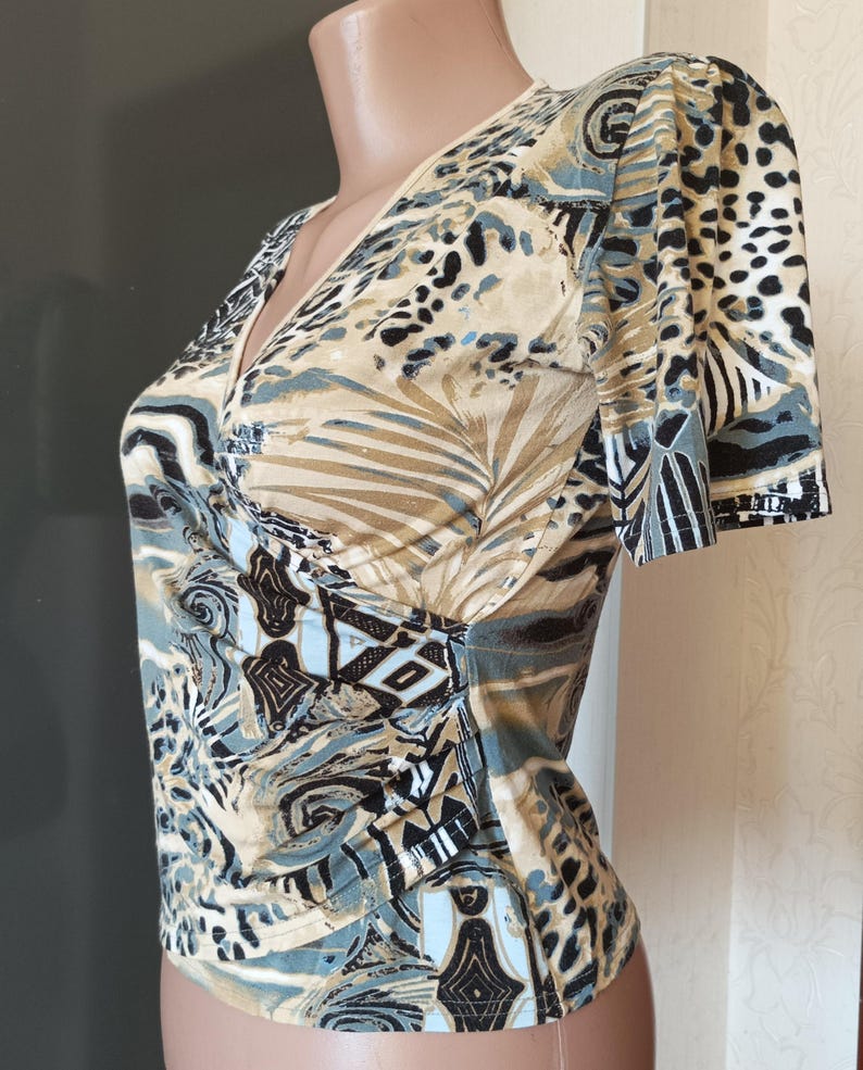 May include: A patterned top with a V-neck and short sleeves. The design features a mix of animal print and abstract shapes in beige, black, and blue. The fabric has a soft appearance. The top has a wrap-style design.