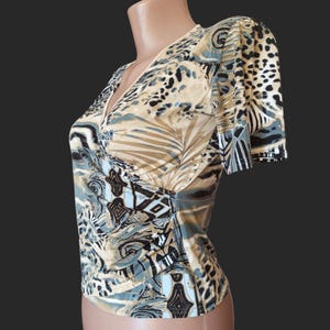 May include: A short-sleeved top with a V-neck and a wrap-style design. The fabric features a pattern with tan, black, and blue abstract shapes, including leaf and animal print designs. The top is displayed on a mannequin.