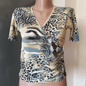 May include: A short-sleeved wrap top with a V-neck. The top has a print with animal and abstract patterns in beige, black, and blue. The fabric looks lightweight and has a slight sheen. The top is a stylish garment.