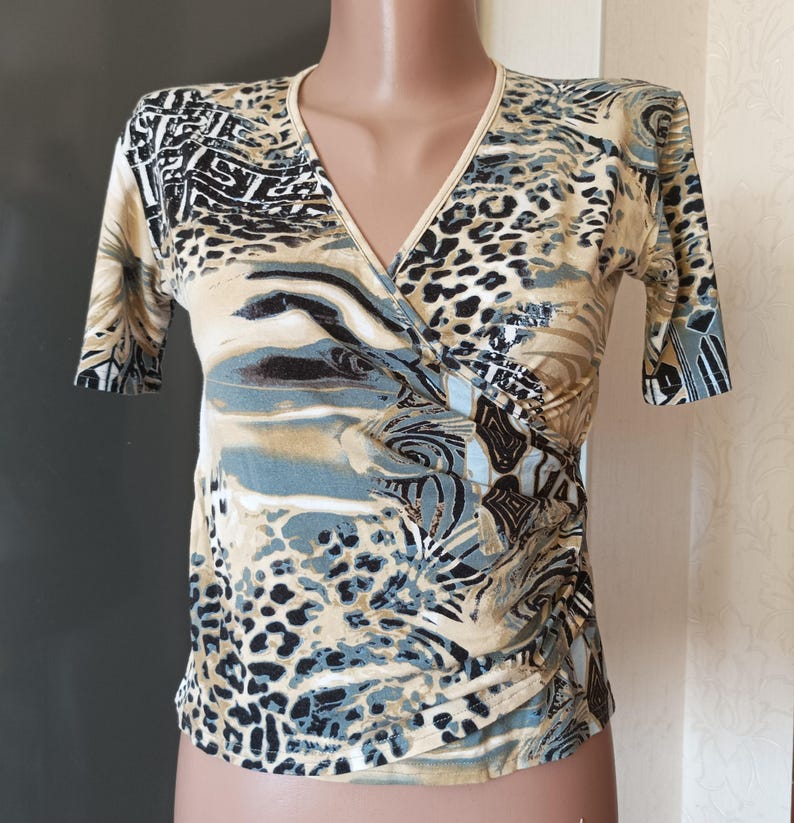 May include: A short-sleeved wrap top with a V-neck. The top has a print with animal and abstract patterns in beige, black, and blue. The fabric looks lightweight and has a slight sheen. The top is a stylish garment.