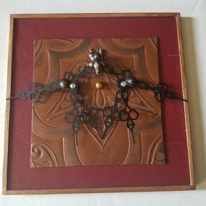 May include: A framed artwork with a brown leather background and a black metal design in the center. The design features a small metal figure and a gold bead.