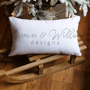 Op de afbeelding: Een wit kussen met de tekst "Eleven & Willow designs" erop, dat op een houten slee ligt.