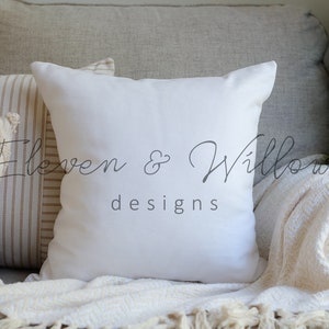 A white cotton-linen pillow sits on a beige couch with a cream chevron-patterned knitted blanket, a white and caramel ticking striped pillow and a beige textured pillow. Christmas tree branches are next to the couch.