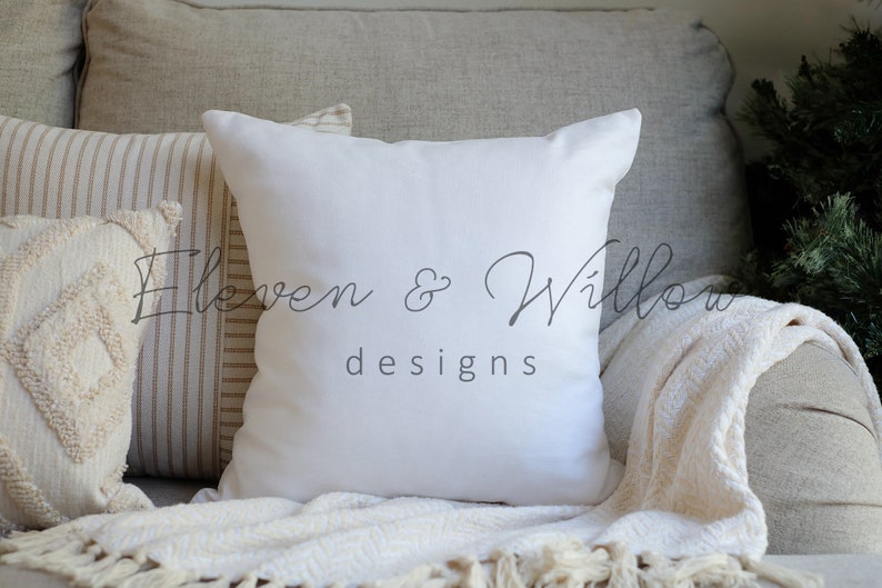 A white cotton-linen pillow sits on a beige couch with a cream chevron-patterned knitted blanket, a white and caramel ticking striped pillow and a beige textured pillow. Christmas tree branches are next to the couch.