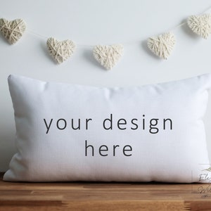 May include: White pillow with the text "your design here" on a wooden surface with a string of white yarn hearts hanging above.