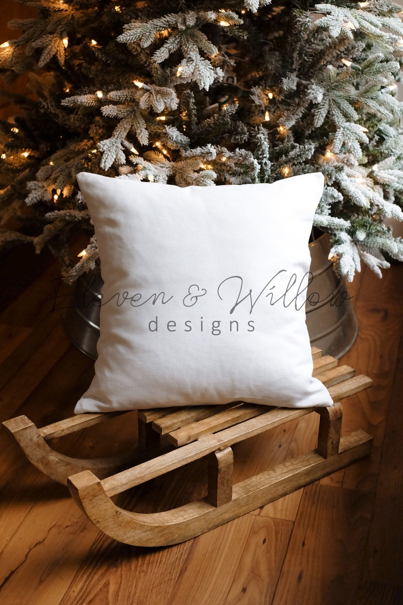 May include: A white pillow with the text "Loven & Willows designs" on a wooden sled in front of a snowy Christmas tree with lights.