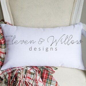 May include: White cushion with the text "Eleven & Willow designs" in black script font. The cushion is sitting on a white chair with a red and green plaid throw blanket.