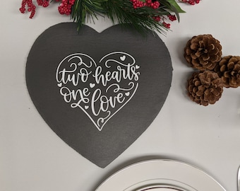 Puede incluir: Un posavasos de pizarra negro en forma de corazón con el texto blanco "two hearts one love" en una fuente cursiva. El posavasos está rodeado de un fondo blanco con piñas y una corona verde con bayas rojas.