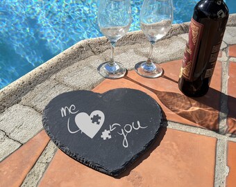 Puede incluir: Un posavasos de pizarra negro en forma de corazón con las palabras "me love you" y un diseño de corazón de pieza de rompecabezas. Dos copas de vino y una botella de vino están sobre una superficie de azulejos junto a una piscina.