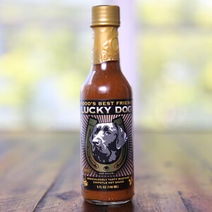 May include: A glass bottle of hot sauce with a gold cap and a black label. The label features a black and white illustration of a dog with a horseshoe and the text "Food's Best Friend Lucky Dog" and "Ridiculously Tasty Mustard Chipotle Hot Sauce". The bottle is 5 fl oz (148 ml).