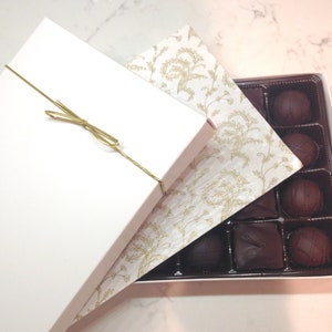 Könnte beinhalten: Eine weiße Schachtel mit einem goldenen Band darum gebunden. Die Schachtel ist geöffnet und innen befindet sich eine Schachtel mit Pralinen. Die Pralinen sind in einem Gittermuster angeordnet und alle aus dunkler Schokolade.