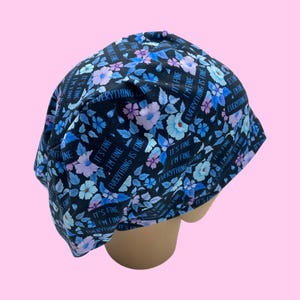 May include: A black scrub cap with a floral pattern in shades of blue and purple. The cap is adorned with the phrases "It's Fine" and "Everything is Fine" in a blue font. The cap is designed to fit the head.