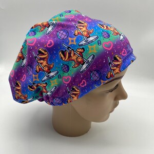 May include: A colorful surgical scrub cap featuring a repeating pattern of orange cartoon dinosaurs pole dancing. The background has a rainbow gradient with hearts, musical notes, and disco balls.
