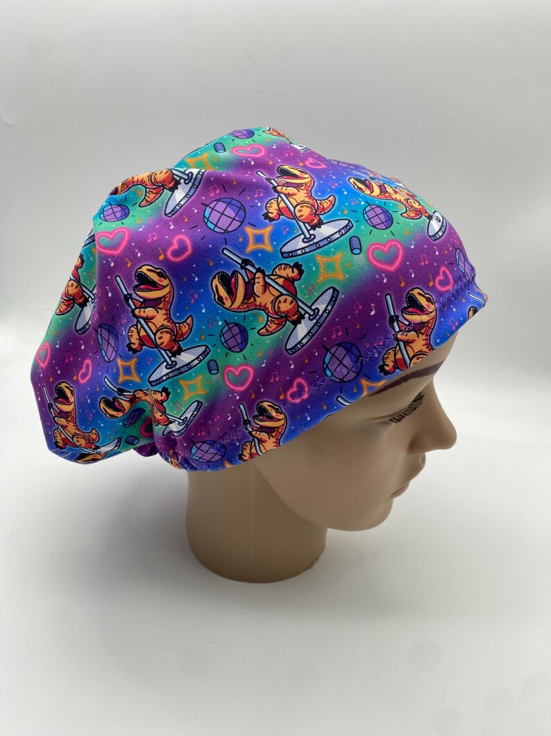 May include: A colorful surgical scrub cap featuring a repeating pattern of orange cartoon dinosaurs pole dancing. The background has a rainbow gradient with hearts, musical notes, and disco balls.
