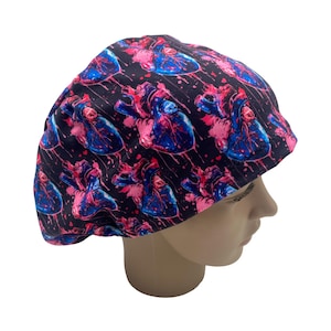 May include: A black scrub cap featuring a repeating pattern of stylized human hearts in shades of pink and blue. The hearts are set against a background of black with pink splatter details. The cap is designed to fit the head.