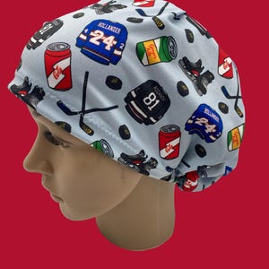Op de afbeelding: Een lichtblauwe operatiemuts met een hockeythema. De muts is bedrukt met hockeytruien, schaatsen, pucks en frisdrankblikjes. De truien hebben de nummers 24 en 81. De frisdrankblikjes zijn rood en groen.