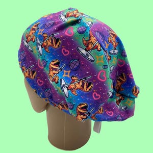 May include: A colorful scrub cap featuring a repeating pattern of cartoon dinosaurs pole dancing. The background has a rainbow gradient with hearts, stars, and disco balls. The cap is designed to fit the head.