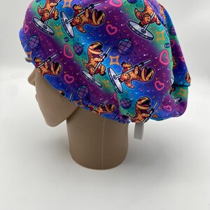 May include: A colorful scrub cap featuring a repeating pattern of cartoon dinosaurs pole dancing. The cap has a vibrant gradient background of purple, blue, and green, with disco balls and hearts. The cap is made of a soft, flexible material.