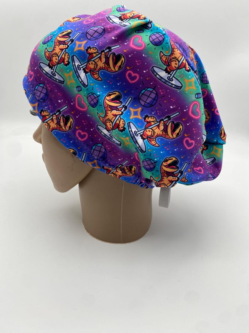 May include: A colorful scrub cap featuring a repeating pattern of cartoon dinosaurs pole dancing. The cap has a vibrant gradient background of purple, blue, and green, with disco balls and hearts. The cap is made of a soft, flexible material.