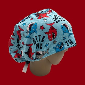 May include: A light blue scrub cap featuring a repeating pattern of cartoon sharks in red, blue, and gray, along with star shapes. The words "Bite Me" are printed on the cap. The cap has a white tie.