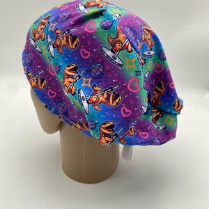 May include: A colorful scrub cap featuring a repeating pattern of cartoon dinosaurs pole dancing. The background has a vibrant gradient of purple, blue, and teal, with hearts, musical notes, and disco balls.